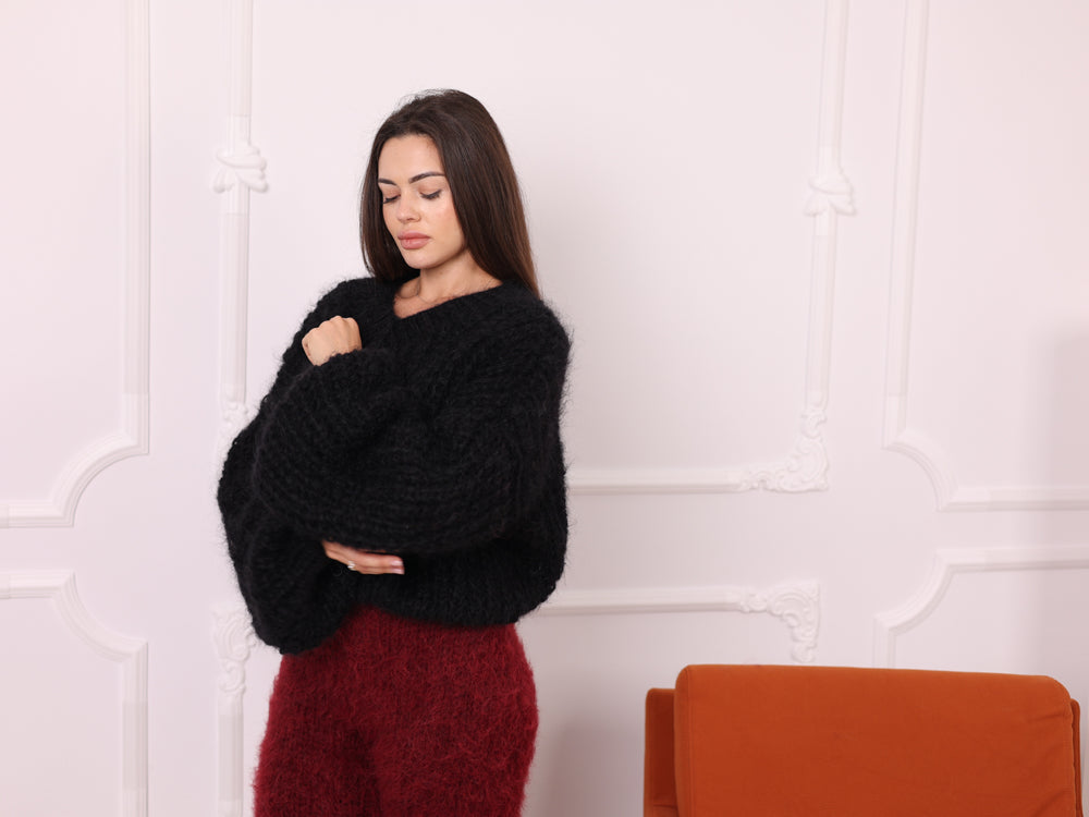 Woman wearing a black sweater and red pants standing against a white wall with an orange chair.