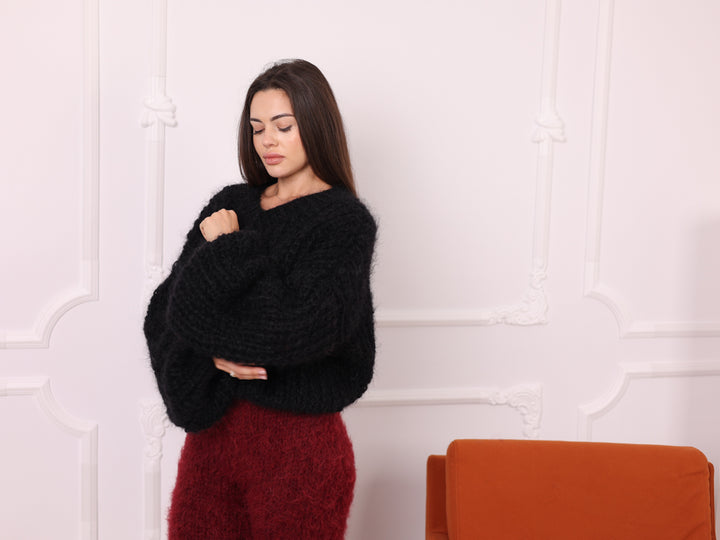 Woman wearing a black sweater and red pants standing against a white wall with an orange chair.