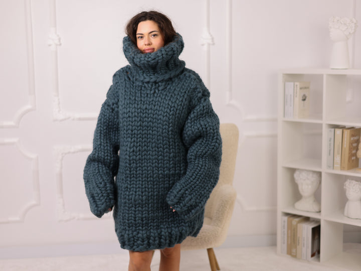Woman wearing a large blue knitted sweater in a room with a white wall and bookshelf.