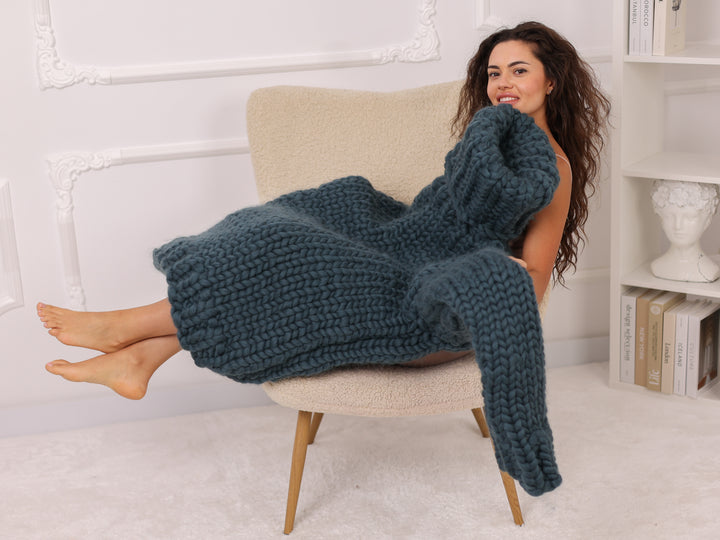 Woman sitting on a chair with a large blue knitted blanket draped over her.