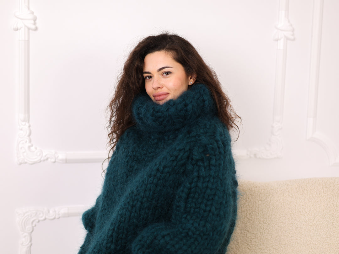 Woman wearing a large teal sweater sitting on a couch against a white wall.