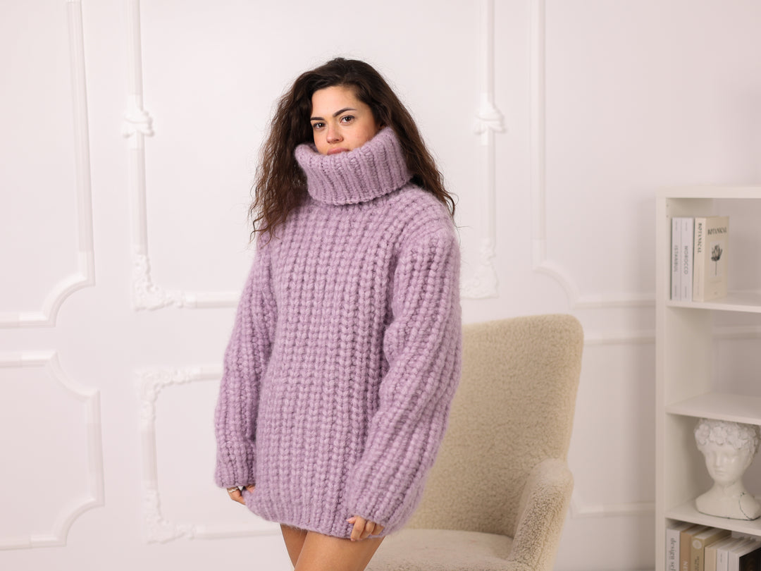 Woman wearing a purple turtleneck sweater in a room with a white wall and a chair.