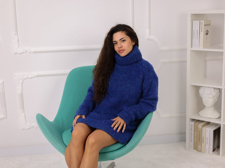 Woman in a blue sweater dress sitting on a green chair against a white wall with a bookshelf.