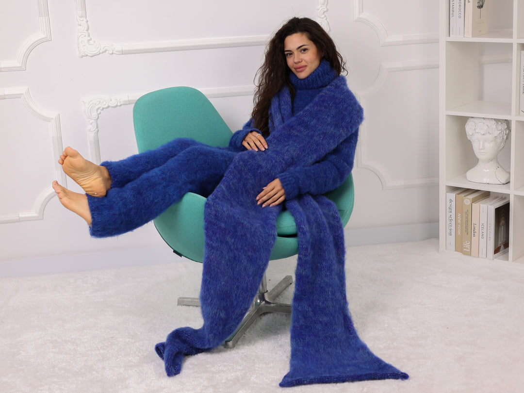 Woman sitting on a chair wrapped in a long blue blanket in a room with a white wall and bookshelf.