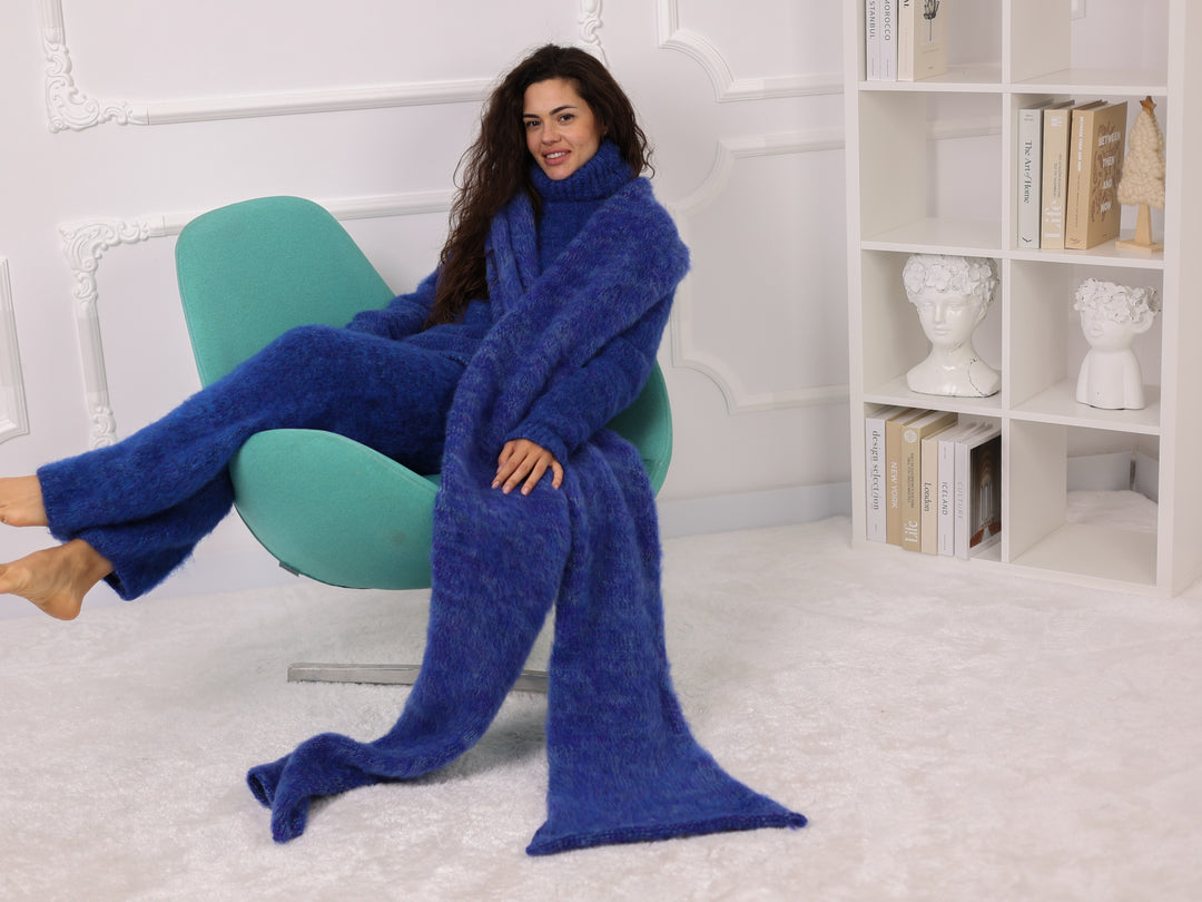 Woman sitting on a green chair wearing a long blue blanket in a room with white walls and a bookshelf.