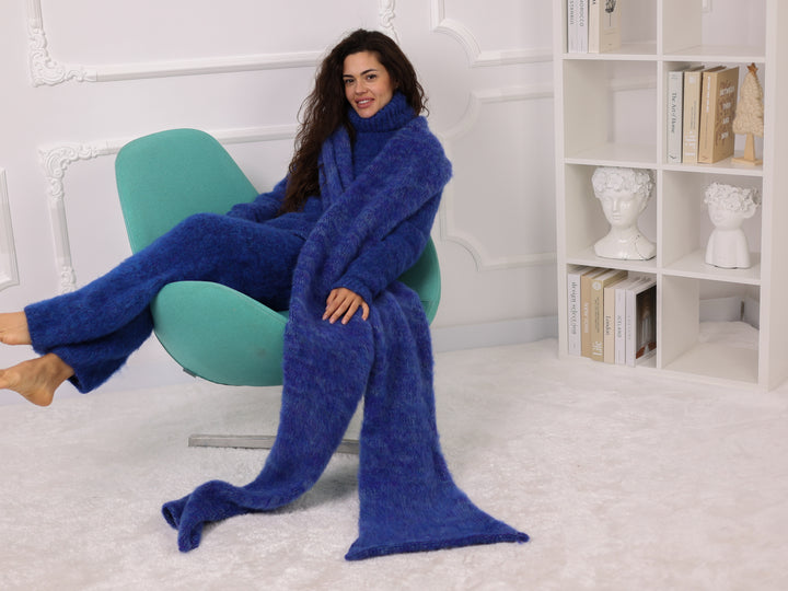 Woman sitting on a green chair wearing a long blue blanket in a room with white walls and a bookshelf.