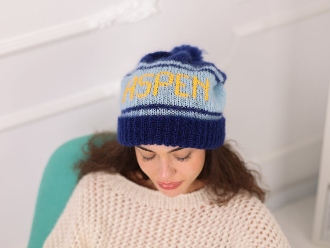 Woman wearing a blue knit hat with 'ASPEM' text, sitting on a white couch.