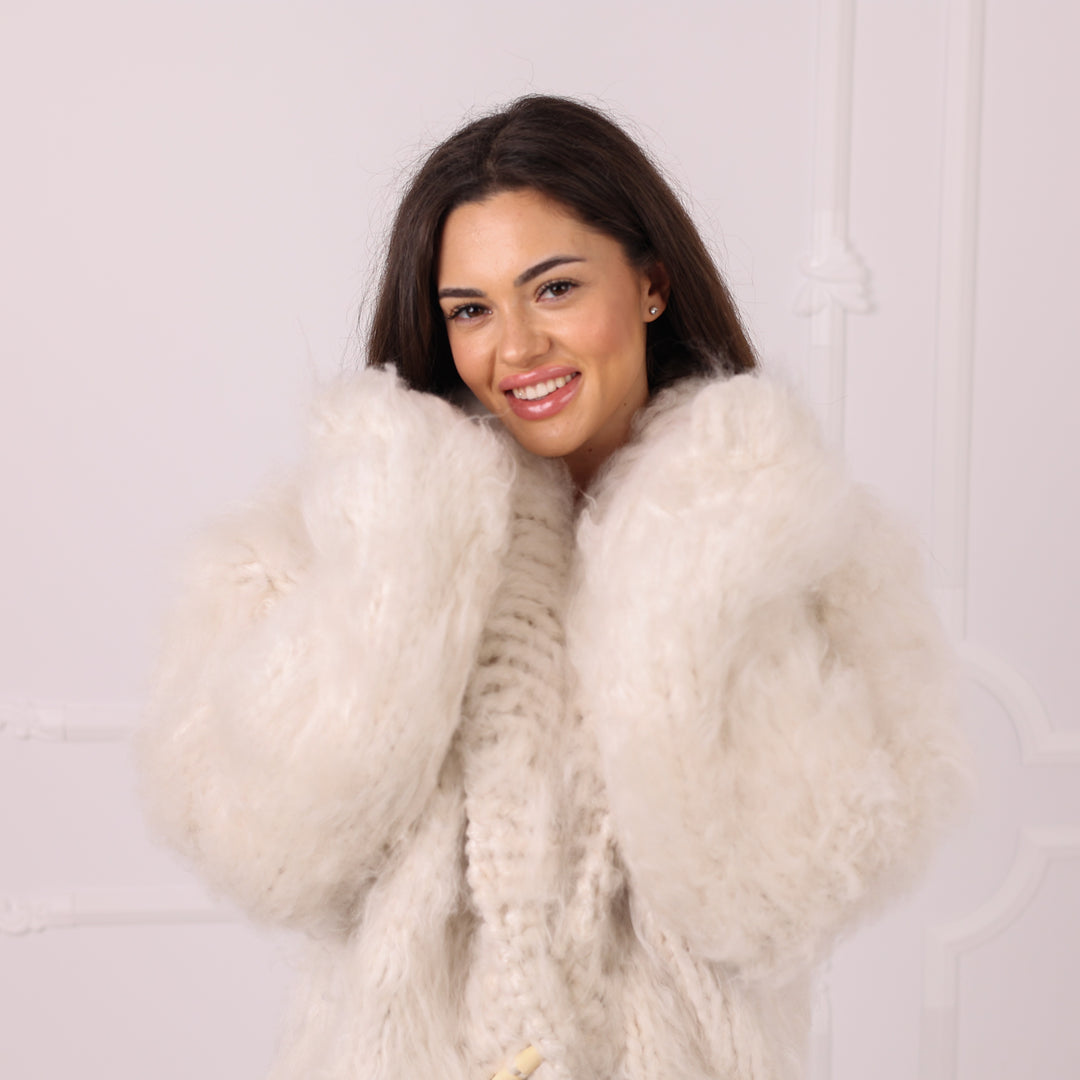 A smiling woman with dark hair wearing a voluminous, fluffy off-white faux fur coat over a cream knit sweater, posing against a light-colored wall with subtle molding details.