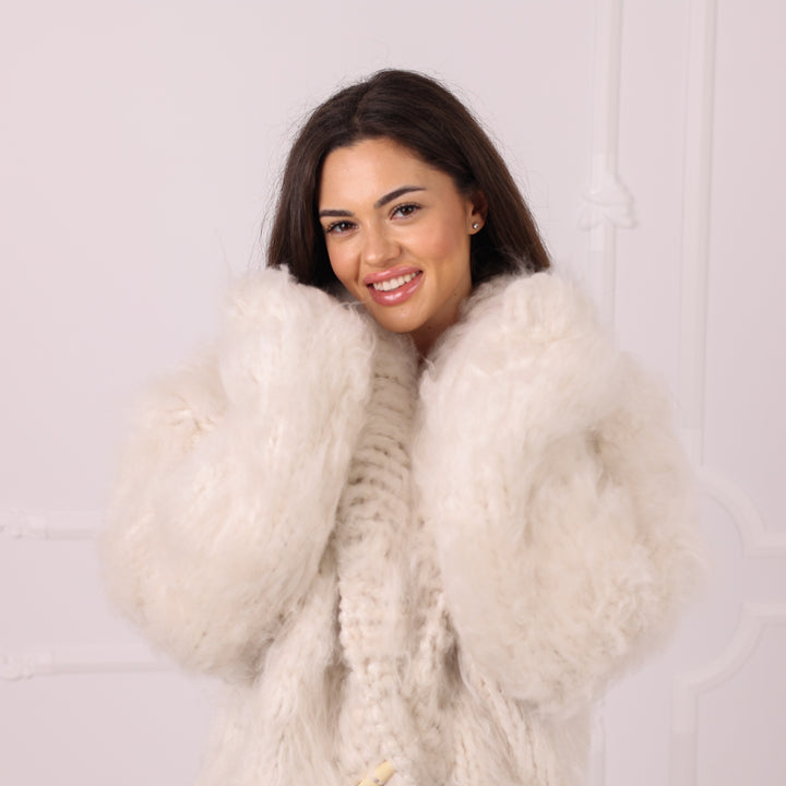 A smiling woman with dark hair wearing a voluminous, fluffy off-white faux fur coat over a cream knit sweater, posing against a light-colored wall with subtle molding details.