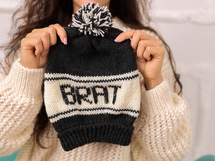 Hand held black and cream soft wool beanie with BRAT text and pom pom