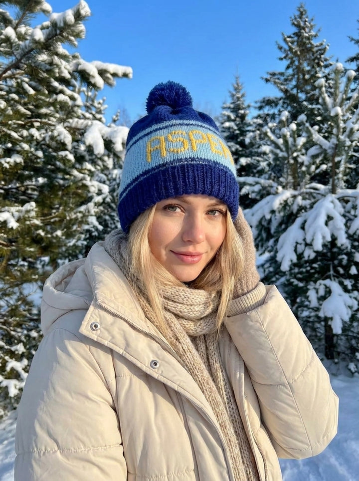 Person wearing a blue knit hat with yellow text and beige coat in a snowy landscape.